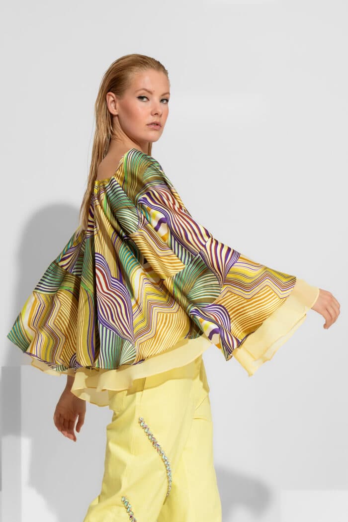 An impressive satin top featuring a vibrant abstract pattern in yellow and green tones. Designed with a sophisticated double flounce and dramatic wide sleeves, this piece offers a voluminous, flowy silhouette. Perfectly paired with high-waisted trousers for a luxurious resort or cocktail look.
